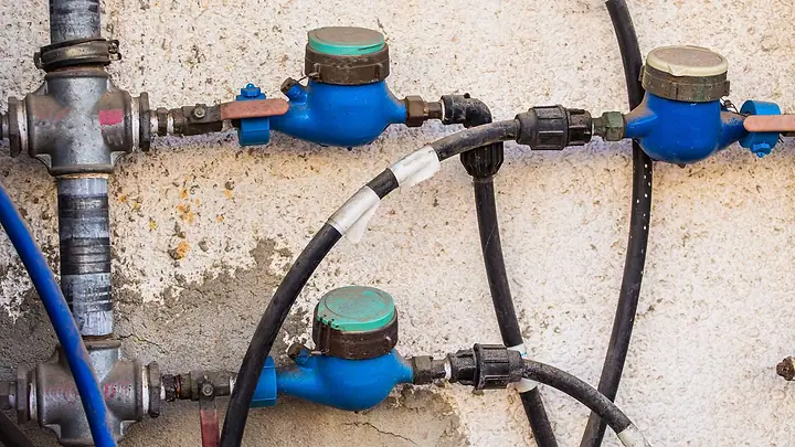 Três hidrômetros azuis instalados numa parede antiga, ligados por canos de metal e mangueiras